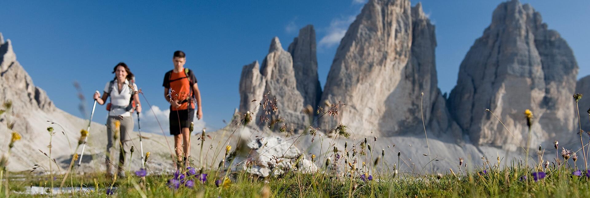 Due escursionisti camminano su un prato fiorito davanti alle Tre Cime sotto un cielo azzurro.