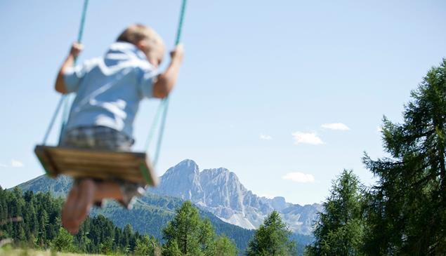Im Vordergrund ein Kind auf einer Schaukel; im Hintergrund die Dolomiten.